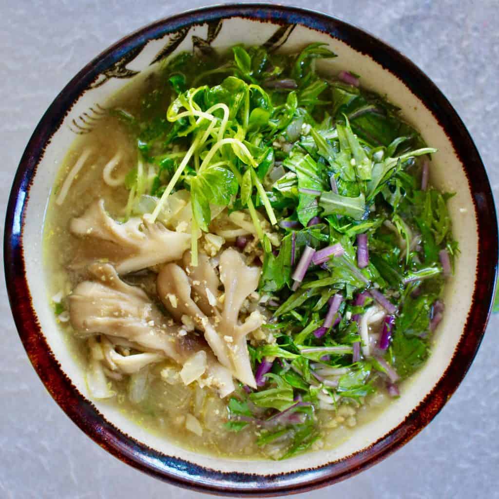 Shirataki noodle ramen soup topped with mushrooms and greens in a light brown bowl with a dark brown rim against a white background