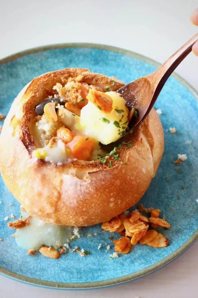 Creamy soup with vegetables and potatoes in a bread bowl on a blue plate with a mouthful being held up with a wooden spoon against a white background