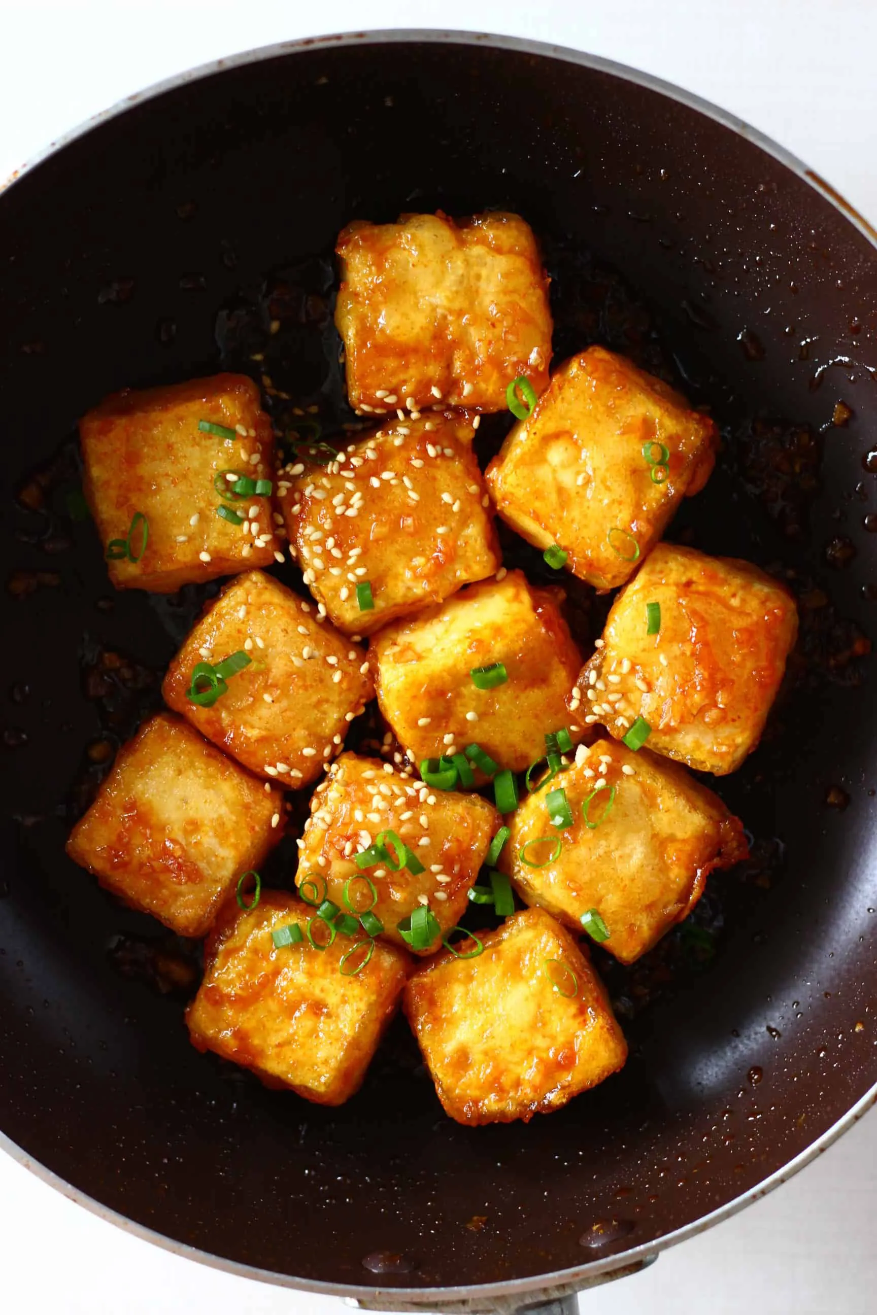 Cubes of spicy tofu sprinkled with sesame seeds and sliced spring onions in a black frying pan