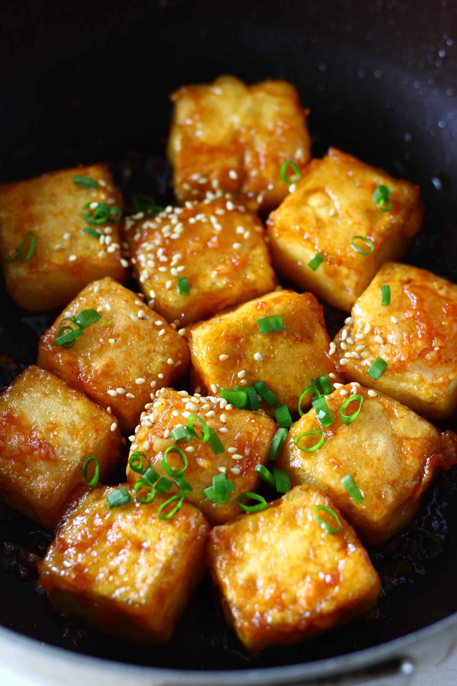 Cubes of spicy tofu sprinkled with sesame seeds and sliced spring onions in a black frying pan