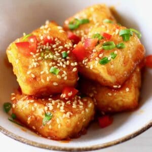 Cubes of spicy tofu sprinkled with sesame seeds, sliced spring onions and chilli in a white bowl with a brown rim against a white background