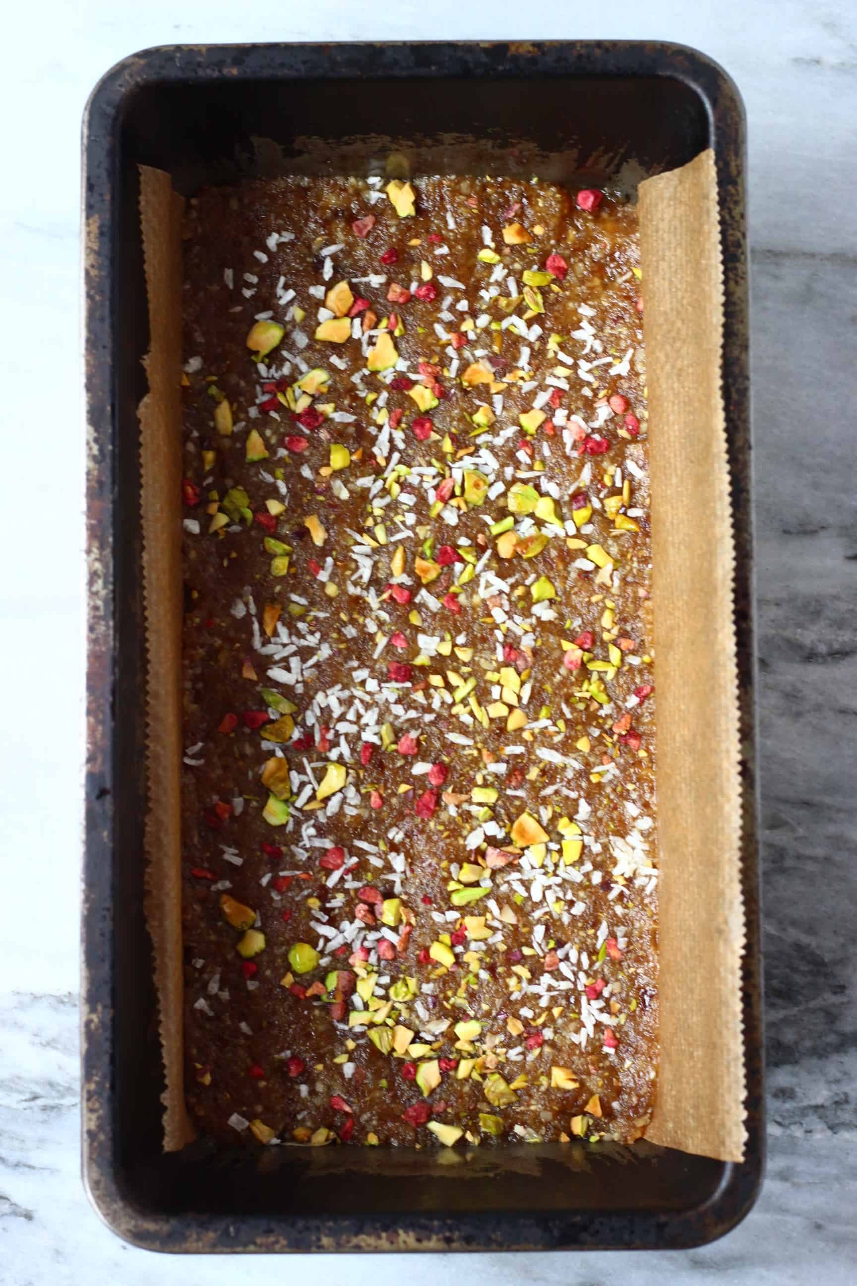 Brown energy bar mixture topped with desiccated coconut, chopped pistachios and freeze-dried raspberries in a loaf tin lined with baking paper