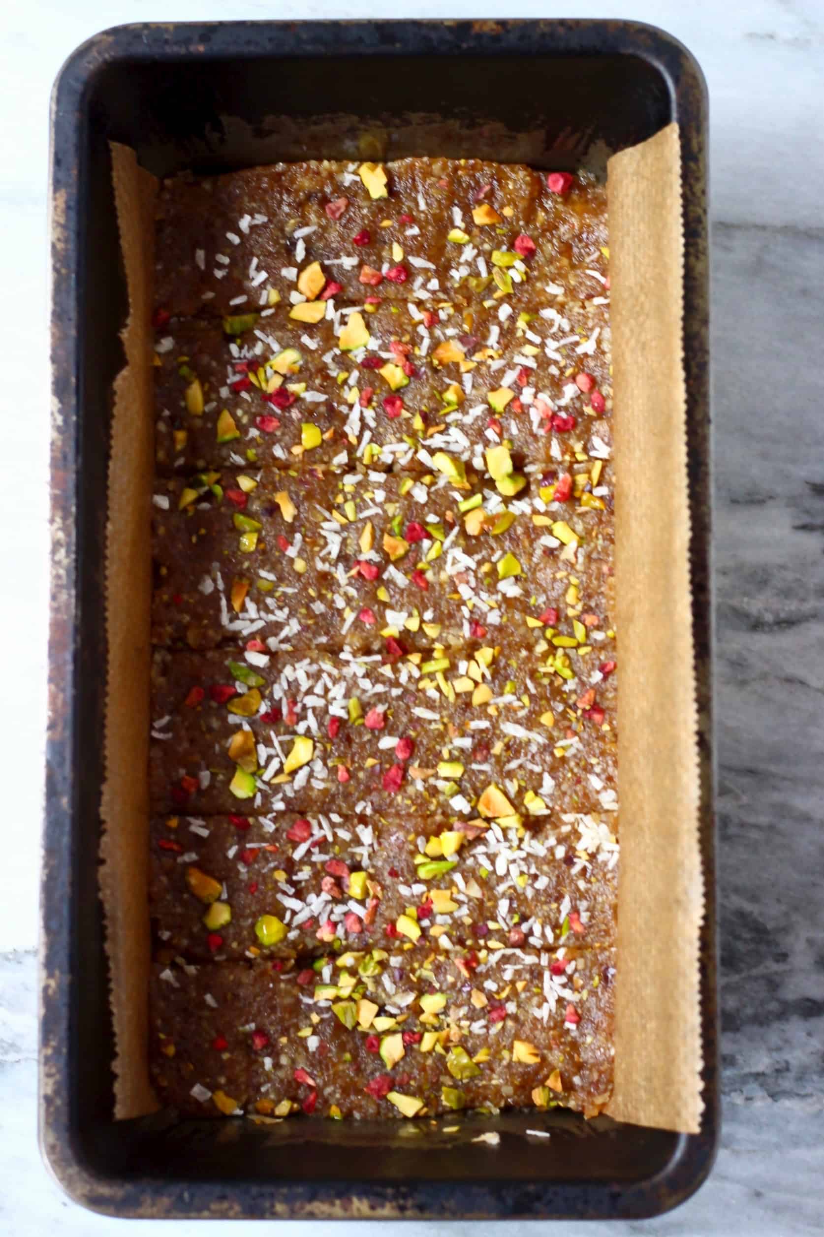 Brown energy bar mixture topped with desiccated coconut, chopped pistachios and freeze-dried raspberries cut into eight pieces in a loaf tin lined with baking paper
