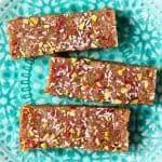 Three brown energy bars sprinkled with chopped pistachio nuts, desiccated coconut and freeze-dried raspberries on a plate