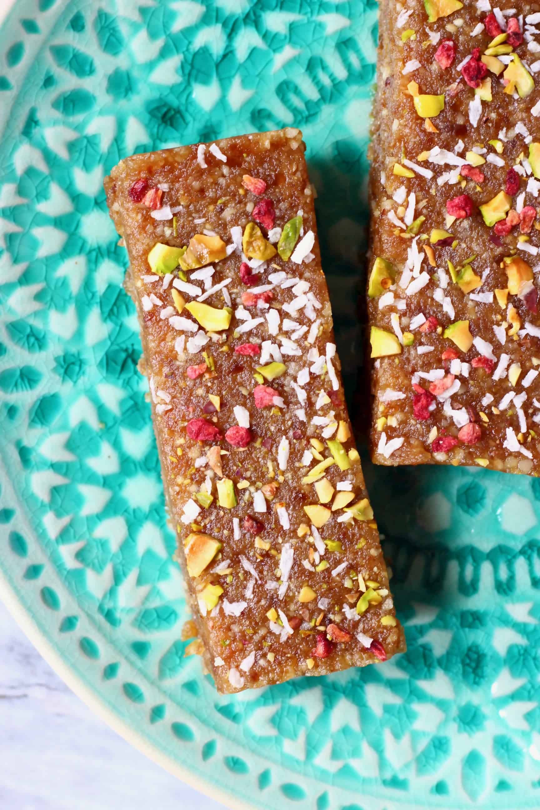 Two brown energy bars sprinkled with chopped pistachio nuts, desiccated coconut and freeze-dried raspberries on a plate