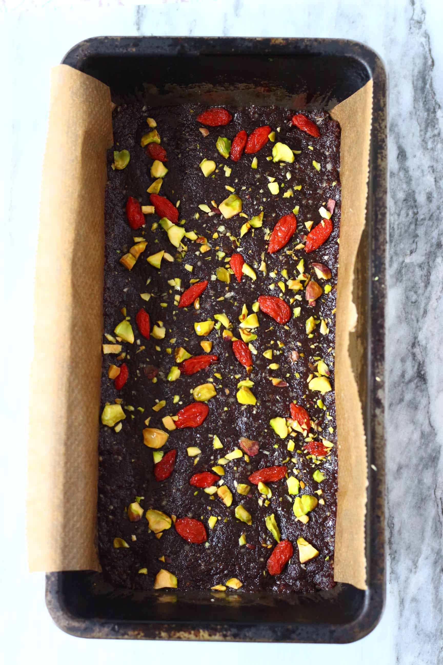 Chocolate no-bake vegan brownie mixture topped with goji berries and chopped pistachios in a loaf tin lined with baking paper
