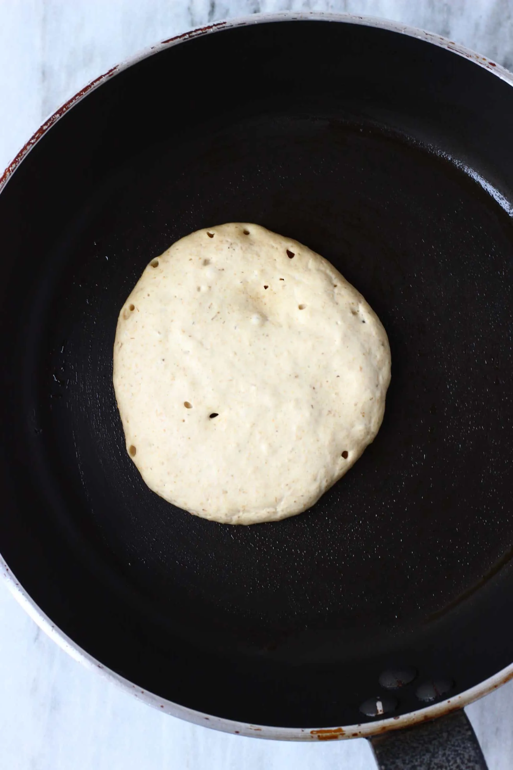 A raw banana oatmeal pancake being cooked in a black frying pan against a marble background
