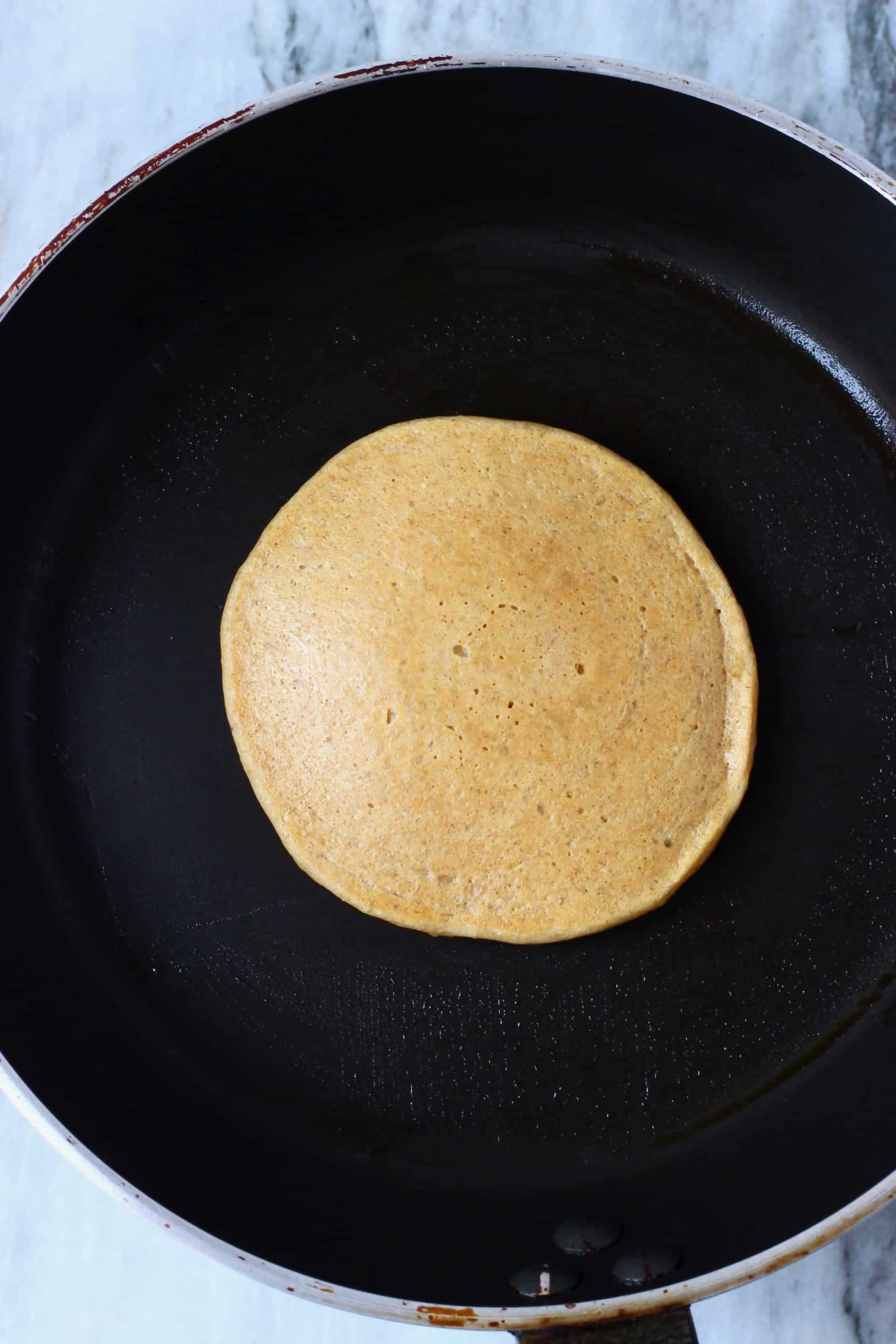 A banana oatmeal pancake being cooked in a black frying pan against a marble background