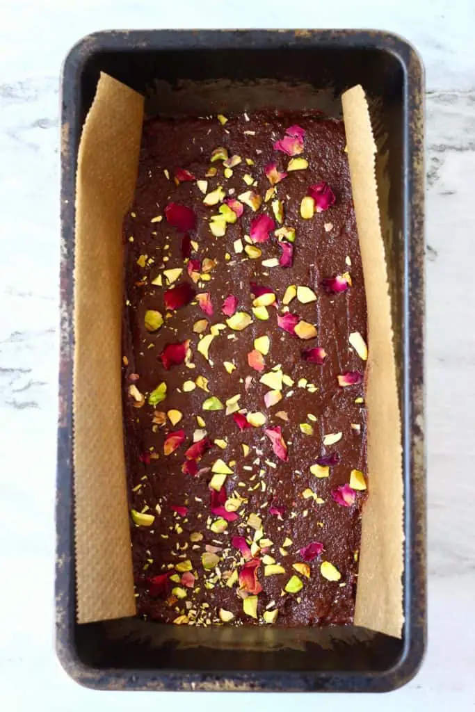 Chocolate bar mixture topped with rose petals and chopped pistachios in a black loaf tin lined with brown baking paper against a marble background