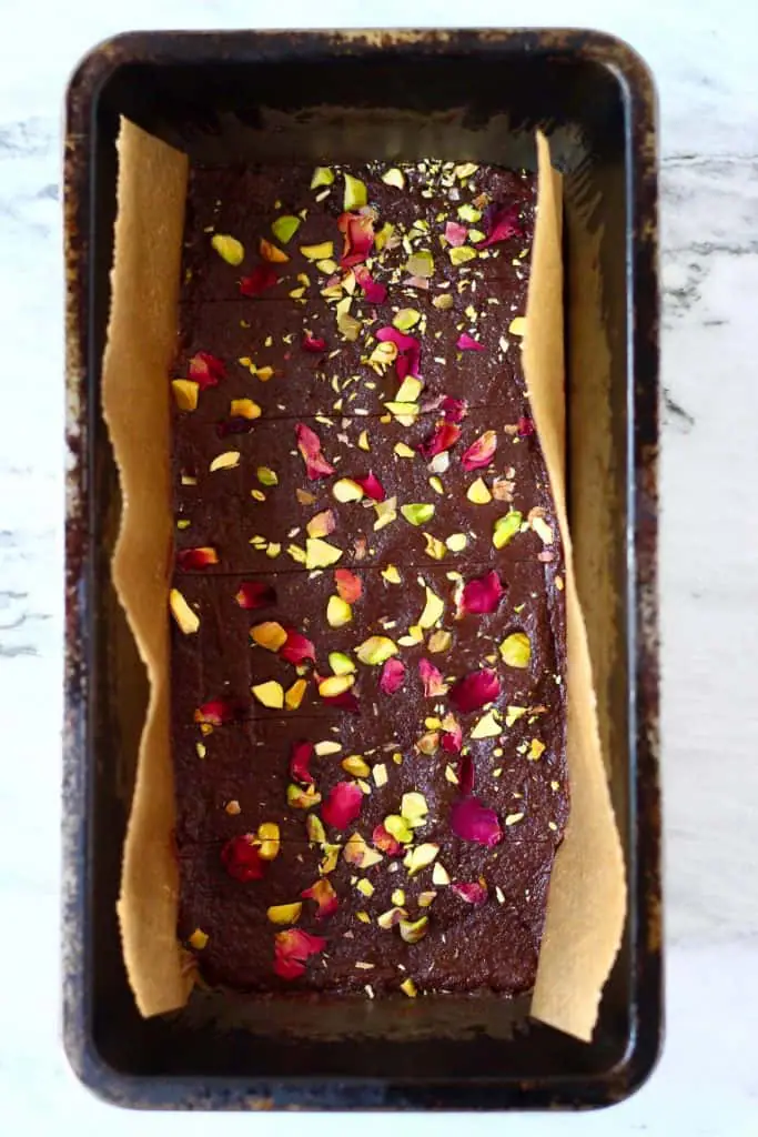 Chocolate bar mixture topped with rose petals and chopped pistachios cut into six horizontal bars in a black loaf tin lined with brown baking paper against a marble background
