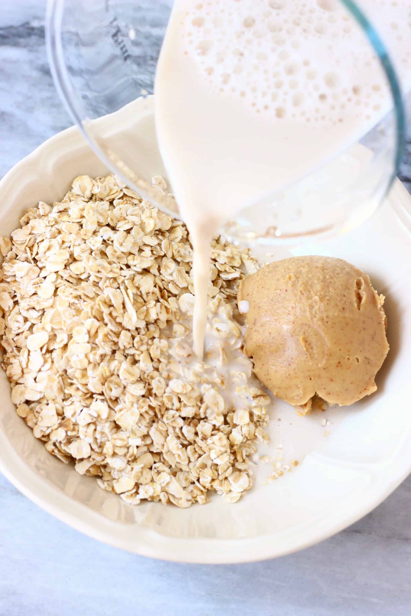 Oats and peanut butter in a white bowl with almond milk in a glass jug being poured in 