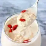 Peanut butter overnight oats in a small glass topped with goji berries with a small silver spoon lifting up a mouthful against a marble background