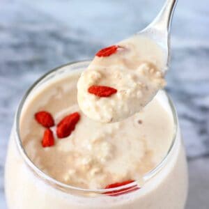 Peanut butter overnight oats in a small glass topped with goji berries with a small silver spoon lifting up a mouthful against a marble background