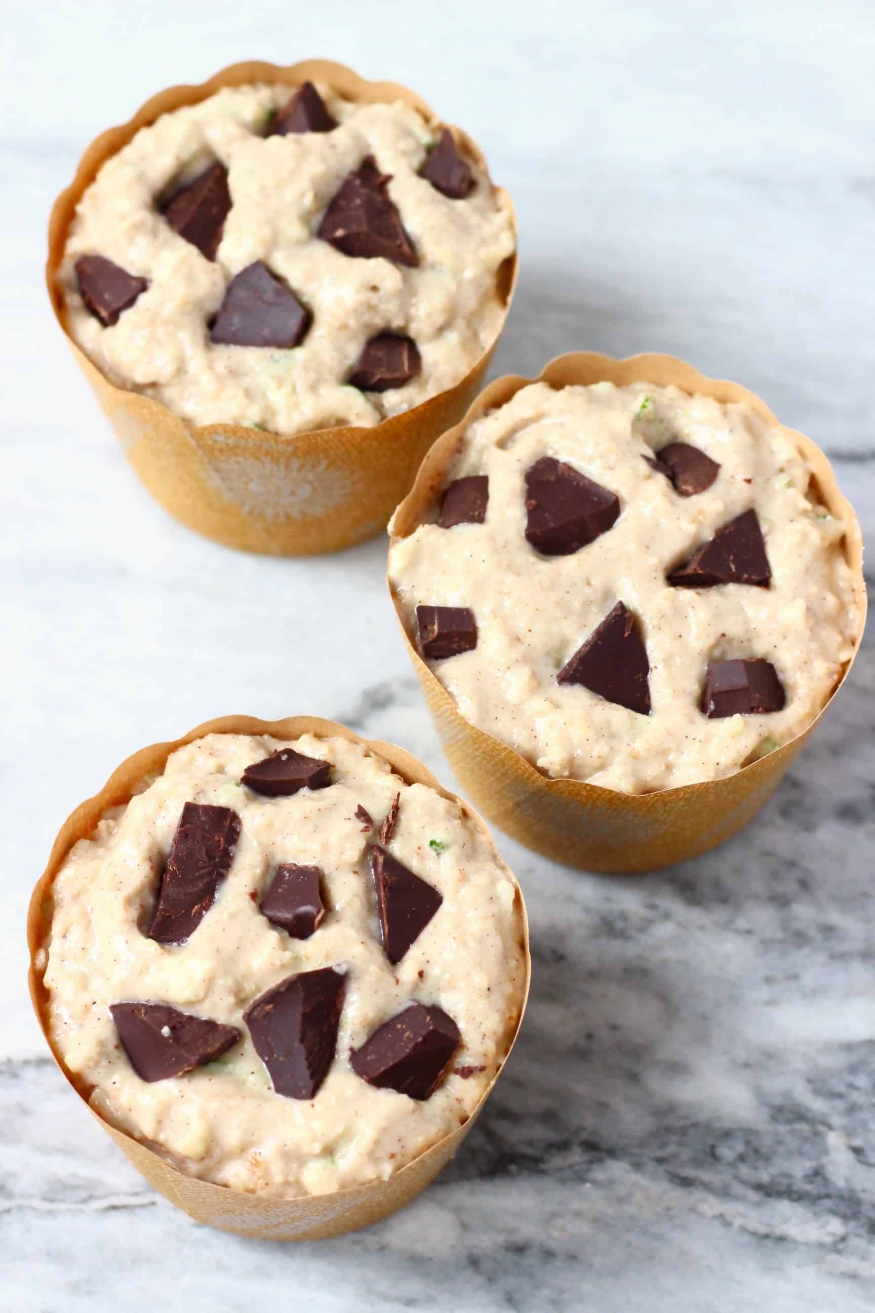 Three brown muffin cases filled with raw zucchini muffin batter with chocolate chunks against a marble background