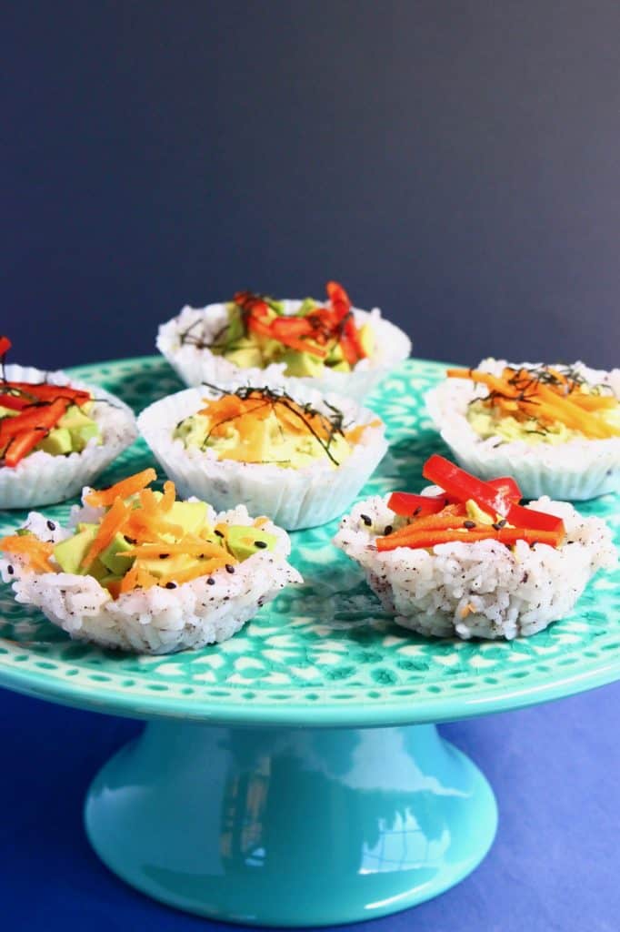 Six muffin-shaped sushi topped with grated carrot and shredded red pepper on a green cake stand against a grey background