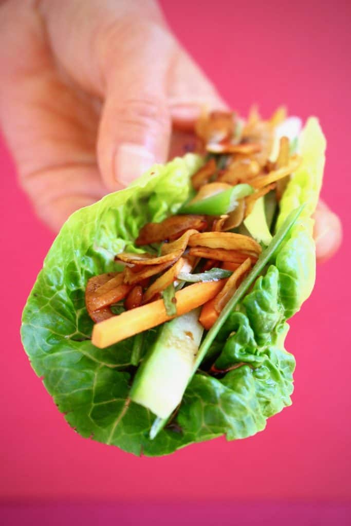 A lettuce wrap filled with brown coconut pieces, sliced spring onions and cucumber held up with a hand against a bright pink background