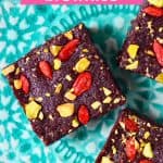 Three square brownies topped with goji berries and chopped pistachios on a green plate