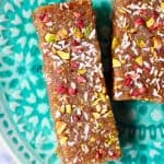 Two brown energy bars sprinkled with chopped pistachio nuts, desiccated coconut and freeze-dried raspberries against a green plate