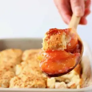 A gluten-free vegan peach cobbler in a grey baking tray with a mouthful being held up by a wooden spoon against a white background
