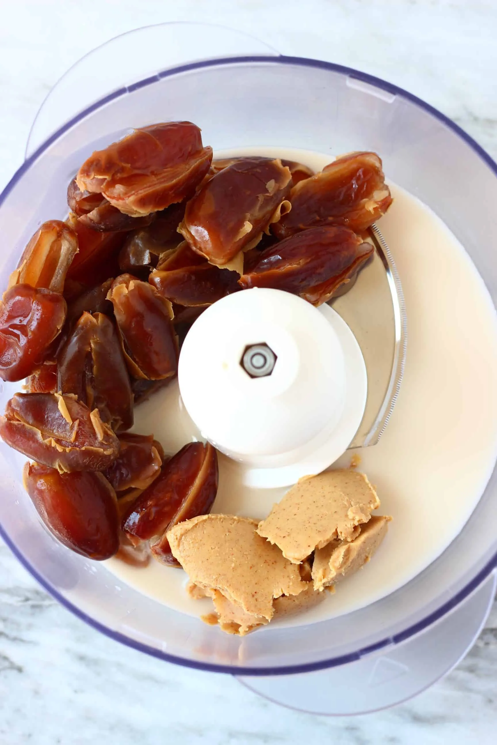 Dates, almond butter and almond milk in a food processor against a marble background