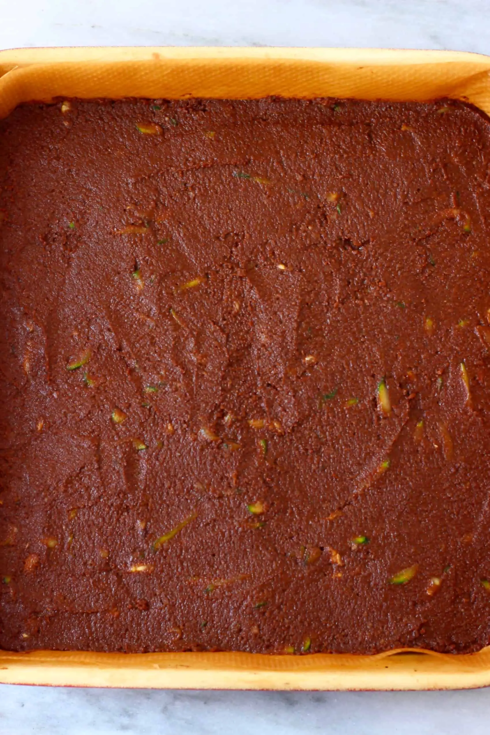 Raw vegan zucchini brownie batter in a square baking tray against a marble background