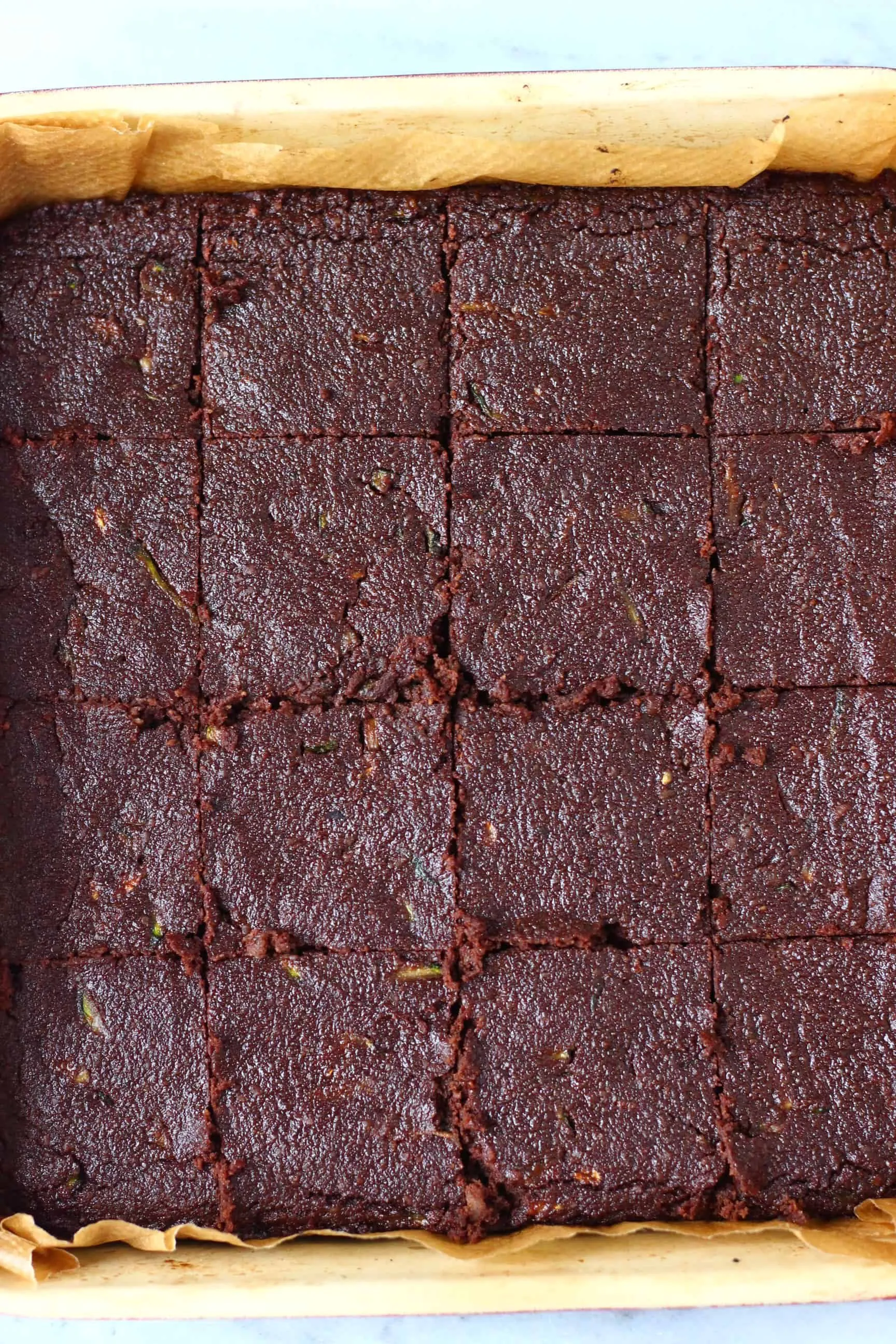 Vegan zucchini brownies in a square baking tray cut into 16 pieces against a marble background