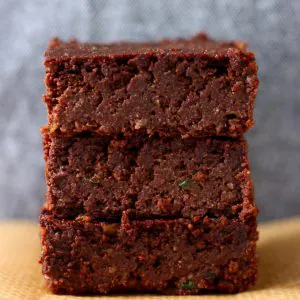 Three vegan zucchini brownies stacked up on top of each other on a sheet of brown baking paper against a grey background