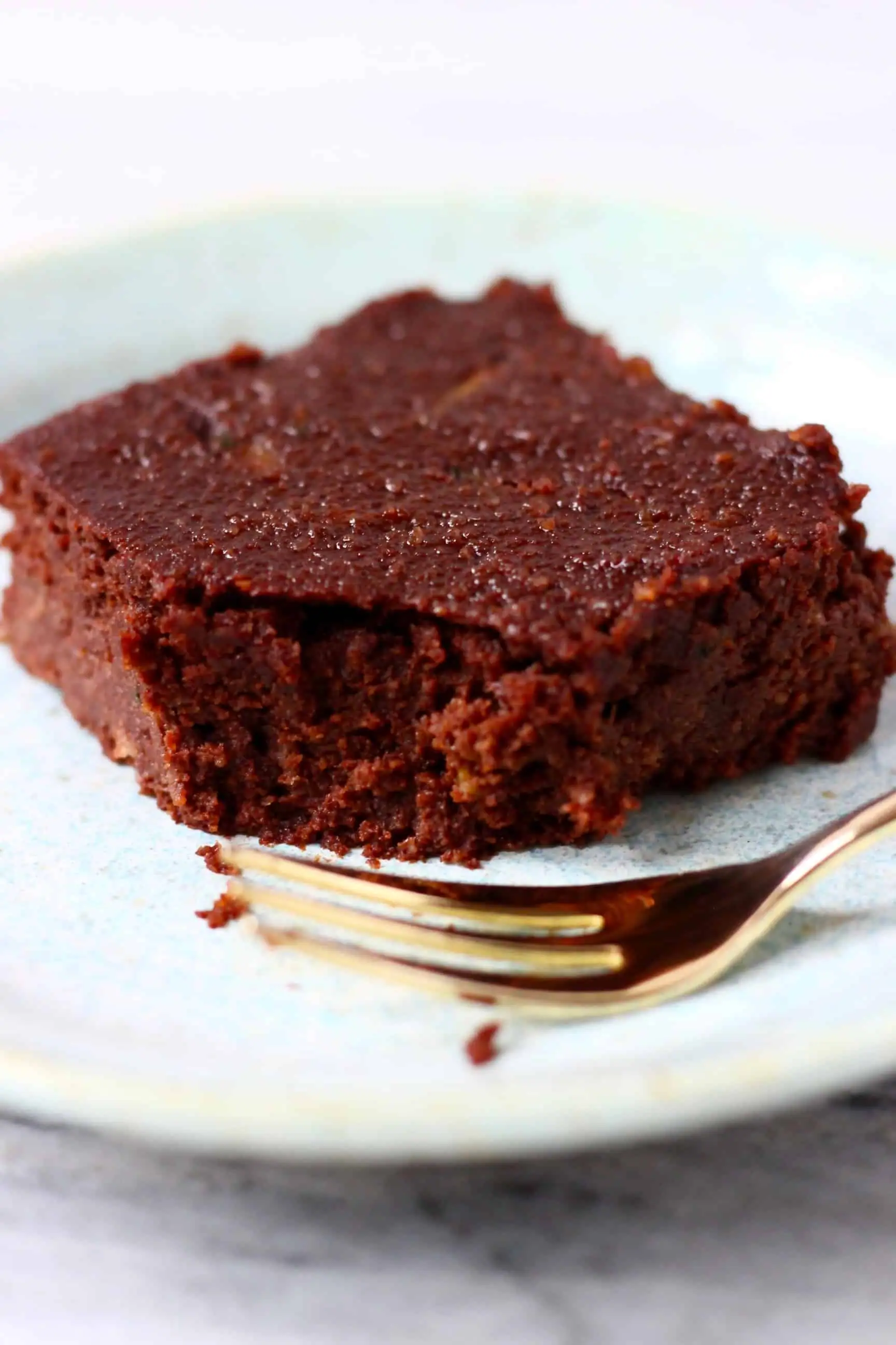 A square vegan zucchini brownie with a bite taken out of it on a blue plate with a gold fork