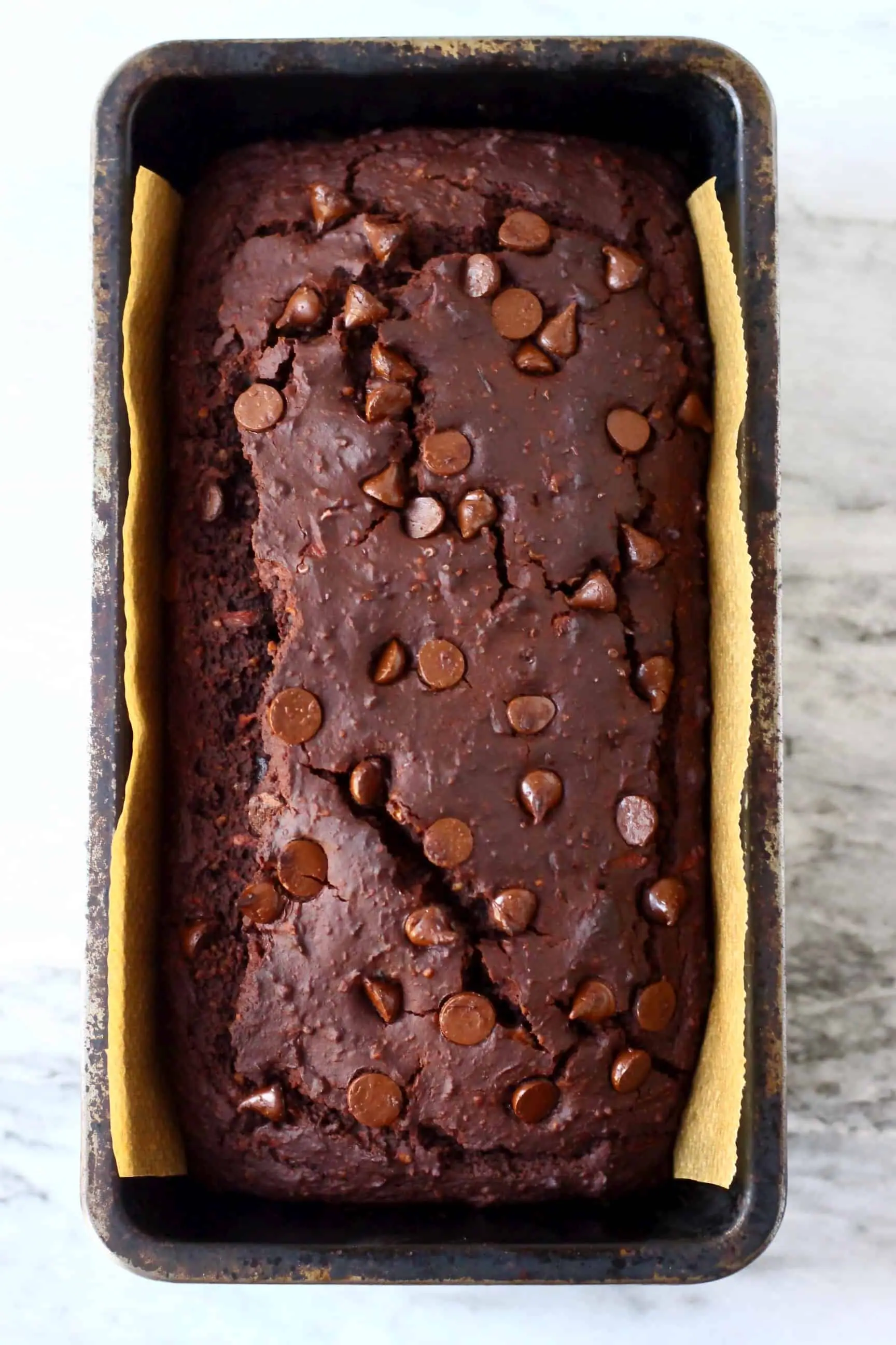 A loaf of gluten-free vegan chocolate zucchini bread topped with chocolate chips in a loaf tin lined with baking paper