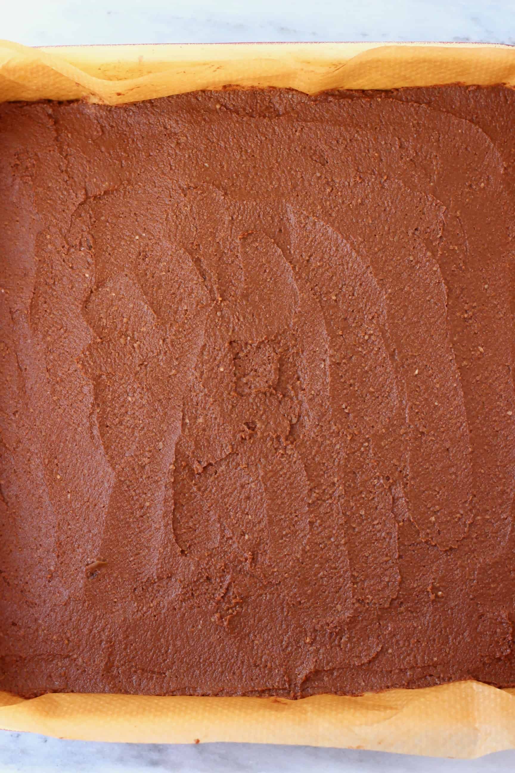 Raw pumpkin brownie batter in a square baking tin lined with brown baking paper against a marble background