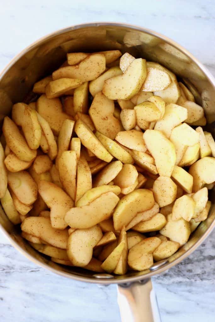 Sliced raw apples with cinnamon and coconut sugar in a silver saucepan against a marble background