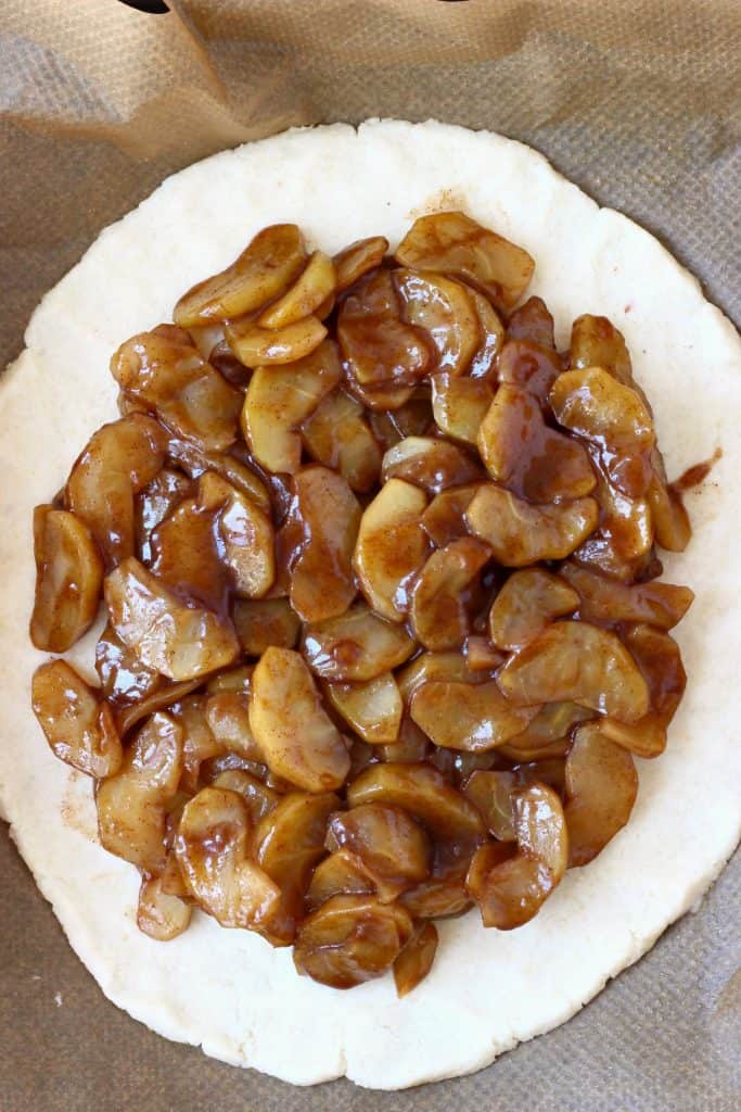 A sheet of raw pastry dough with brown cooked apples in the middle against a sheet of brown baking paper