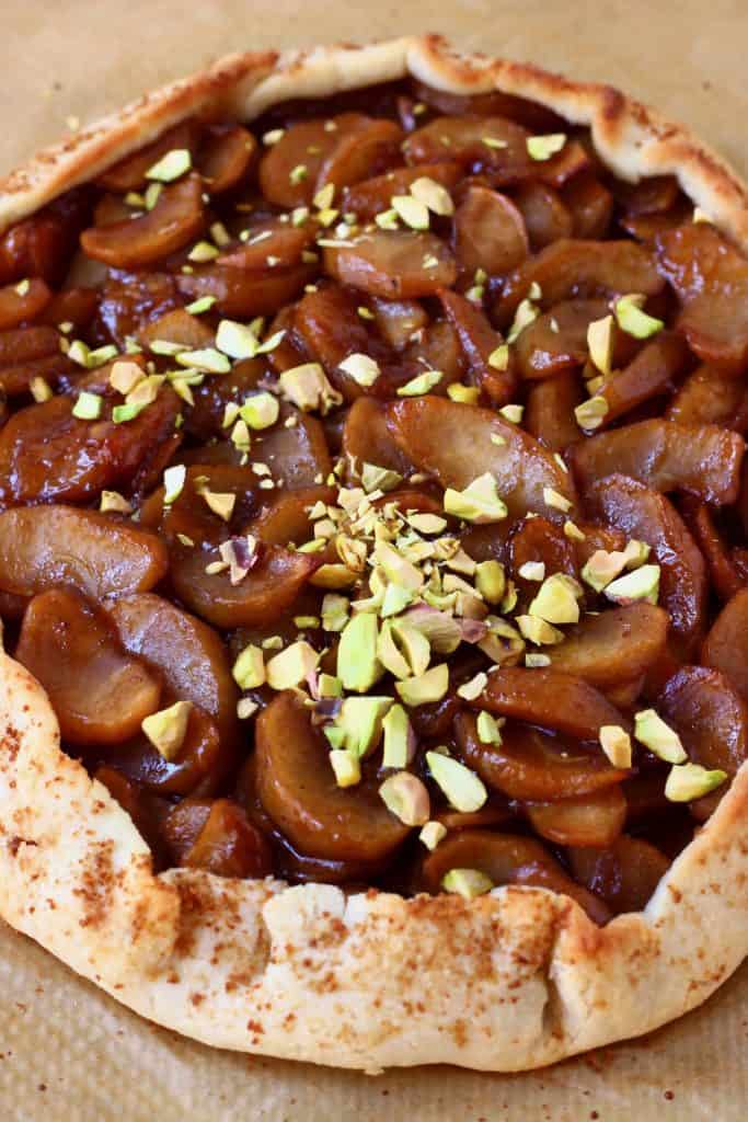 A round galette filled with brown apples sprinkled with pistachio nuts against a sheet of brown baking paper