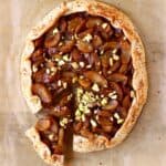 A round galette filled with brown apples sprinkled with pistachio nuts with a slice cut out of it against a sheet of brown baking paper
