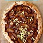 A round galette filled with brown apples sprinkled with pistachio nuts against a sheet of brown baking paper