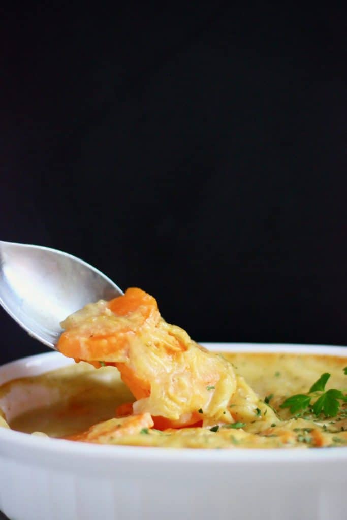 Scalloped sweet potatoes in a white pie dish with a silver spoon lifting up a mouthful against a black background
