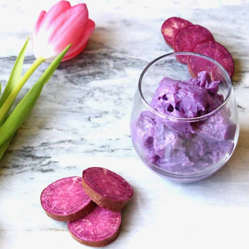 Purple sweet potato ice cream in a glass with slices of raw purple sweet potato and a pink tulip against a marble background