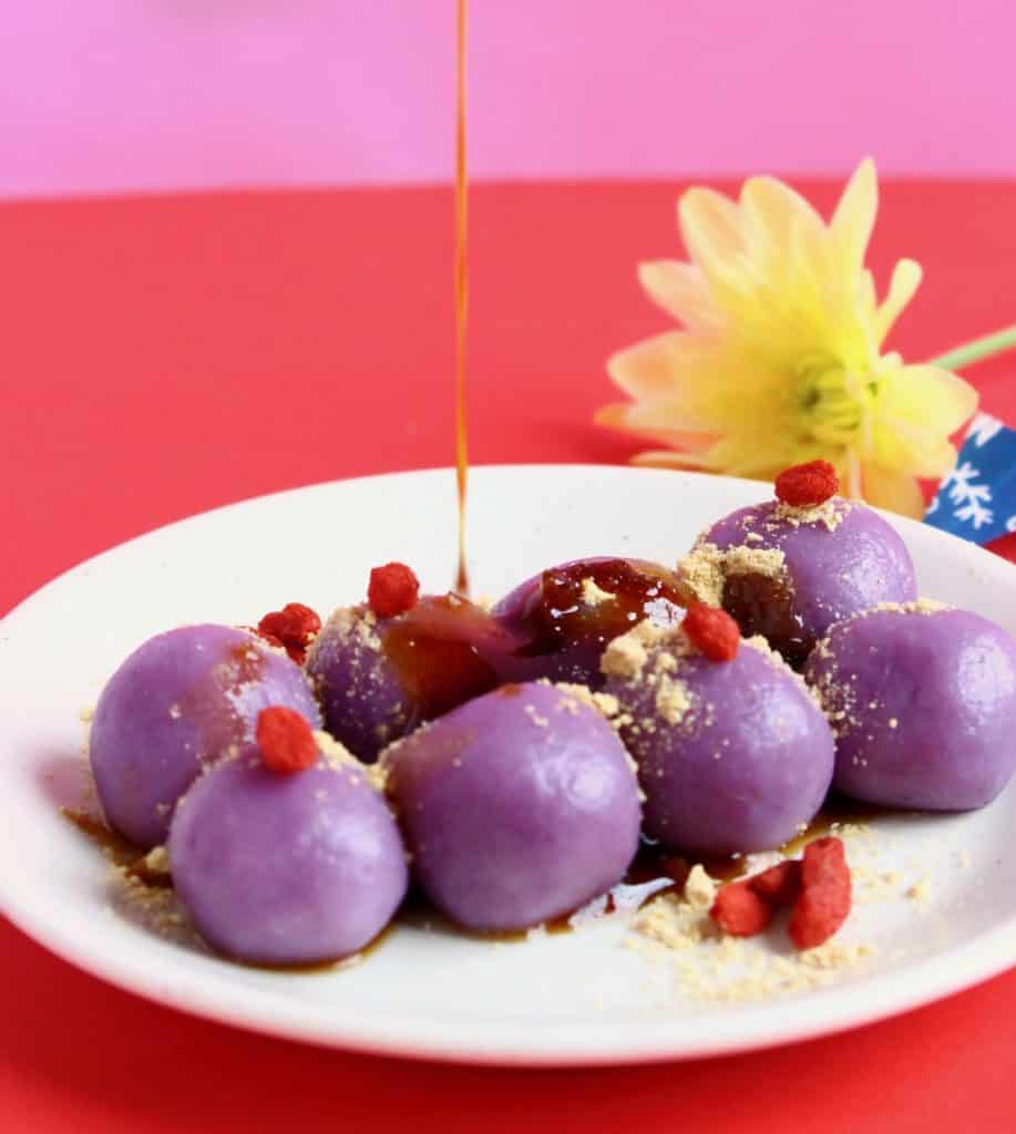 Eight balls of purple sweet potato mochi on a white plate with brown syrup being poured over the top against a pink and red background