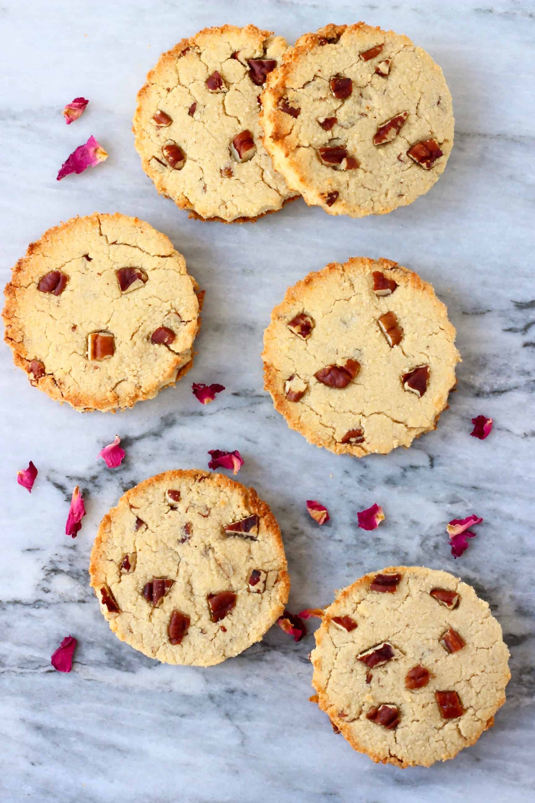 Six round coconut flour cookies with pecan nuts against a marble background scattered with rose petals