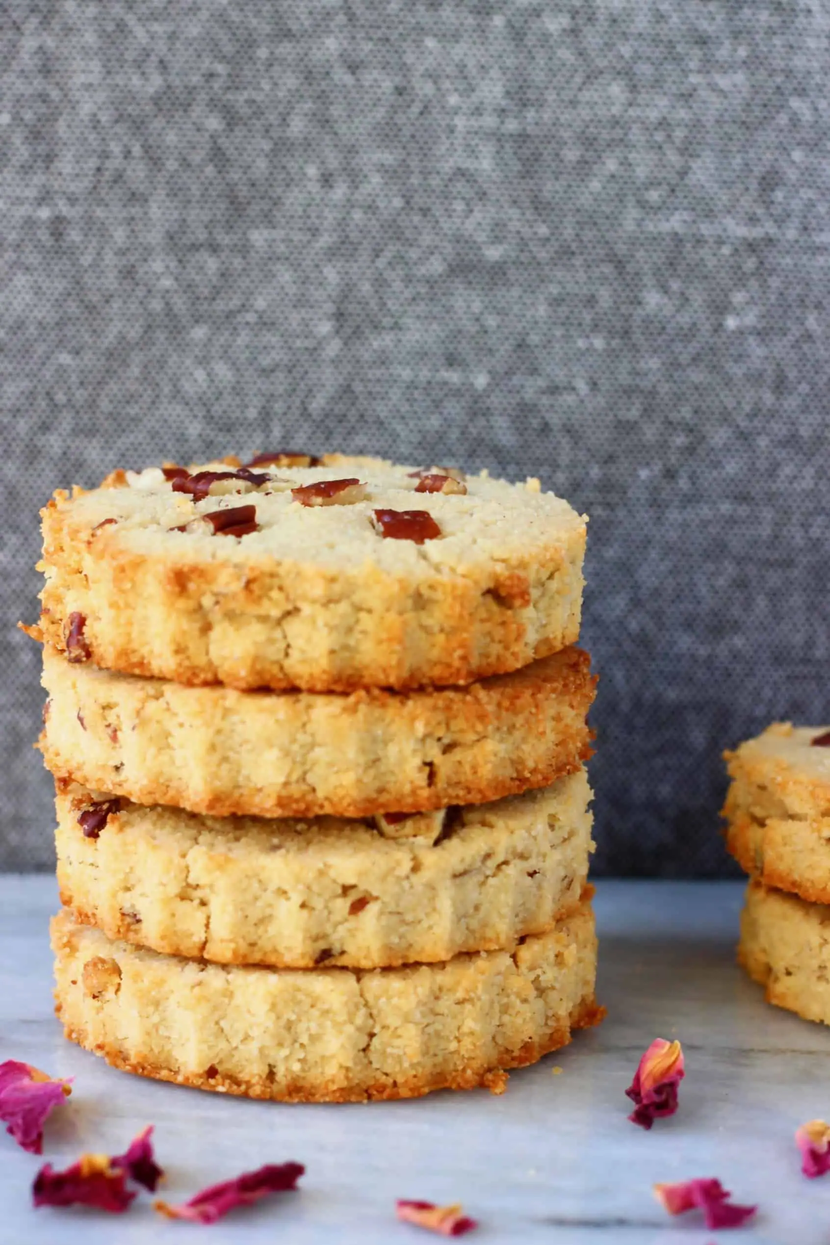 Four circular coconut flour cookies with pecan nuts stacked up on a marble slab against a grey background scattered with rose petals