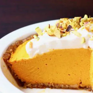 A white pie dish with no-bake vegan pumpkin pie topped with whipped cream and chopped pistachios against a dark brown background