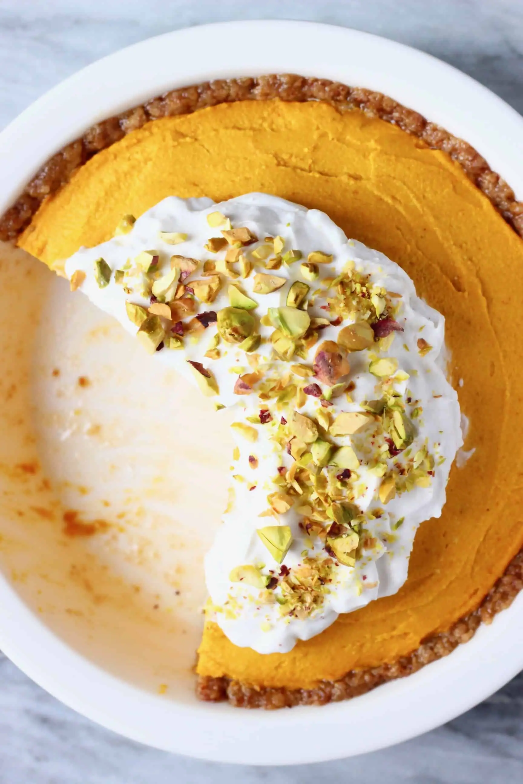No-bake vegan pumpkin pie topped with whipped cream and chopped pistachios with a slice taken out of it in a white pie dish