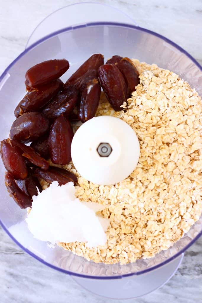 Dates, oats and coconut oil in a food processor against a marble background