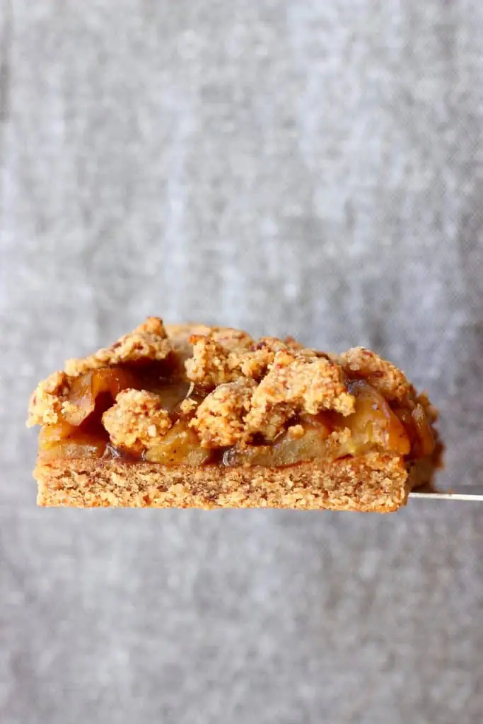 A square apple crumble bar held up with a silver spatula against a grey background