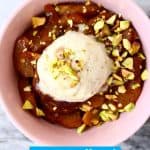 Golden brown baked apples in a pink bowl scattered with chopped pistachio nuts and topped with vanilla ice cream against a marble background