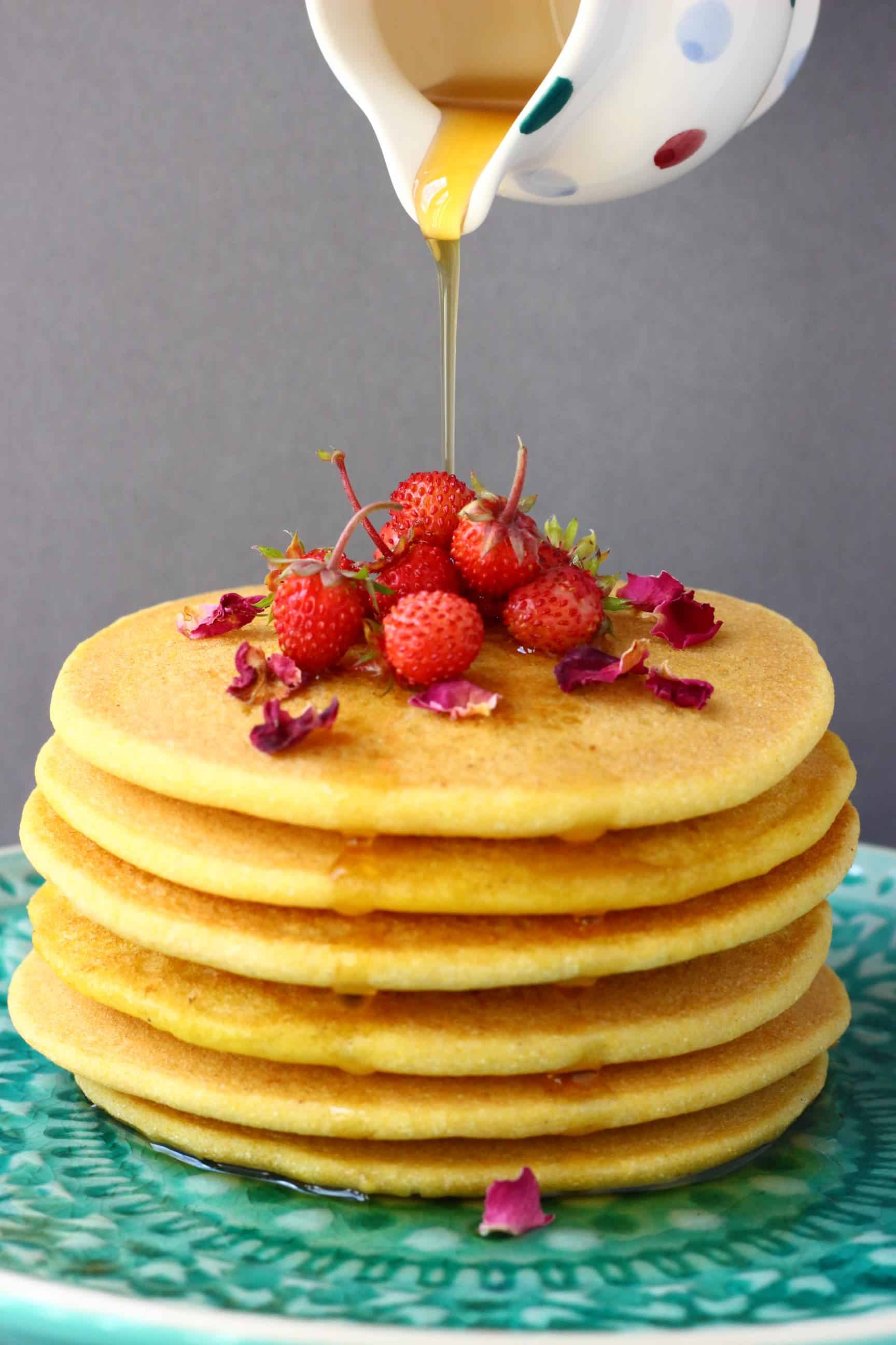 A stack of six gluten-free vegan cornmeal pancakes on a plate topped with mini strawberries and rose petals with a jug pouring maple syrup over them