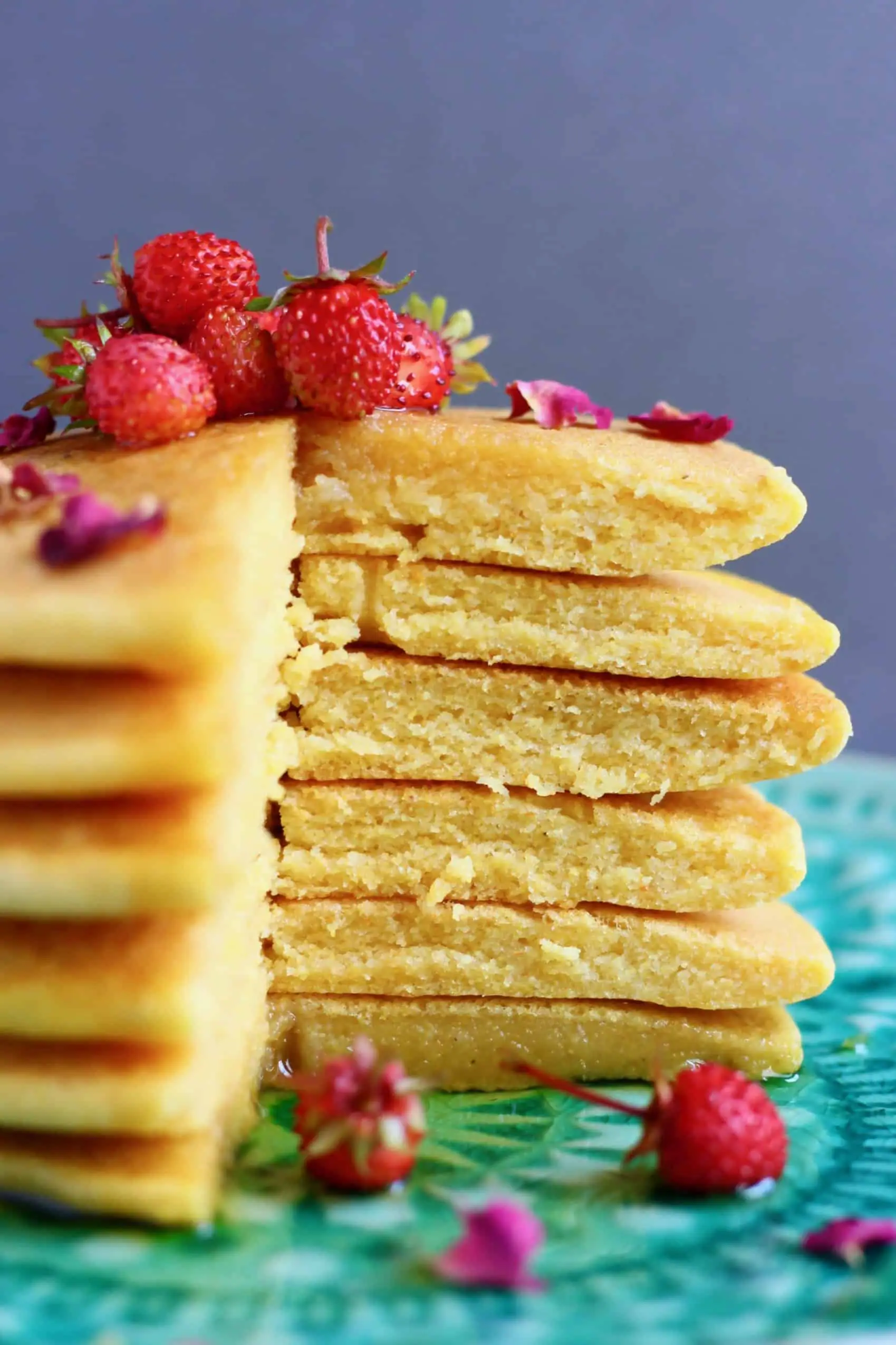A stack of gluten-free vegan cornmeal pancakes with a slice cut out of them topped with rose petals and small strawberries on a plate