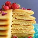 A stack of gluten-free vegan cornmeal pancakes with a slice cut out of them topped with rose petals and small strawberries on a plate