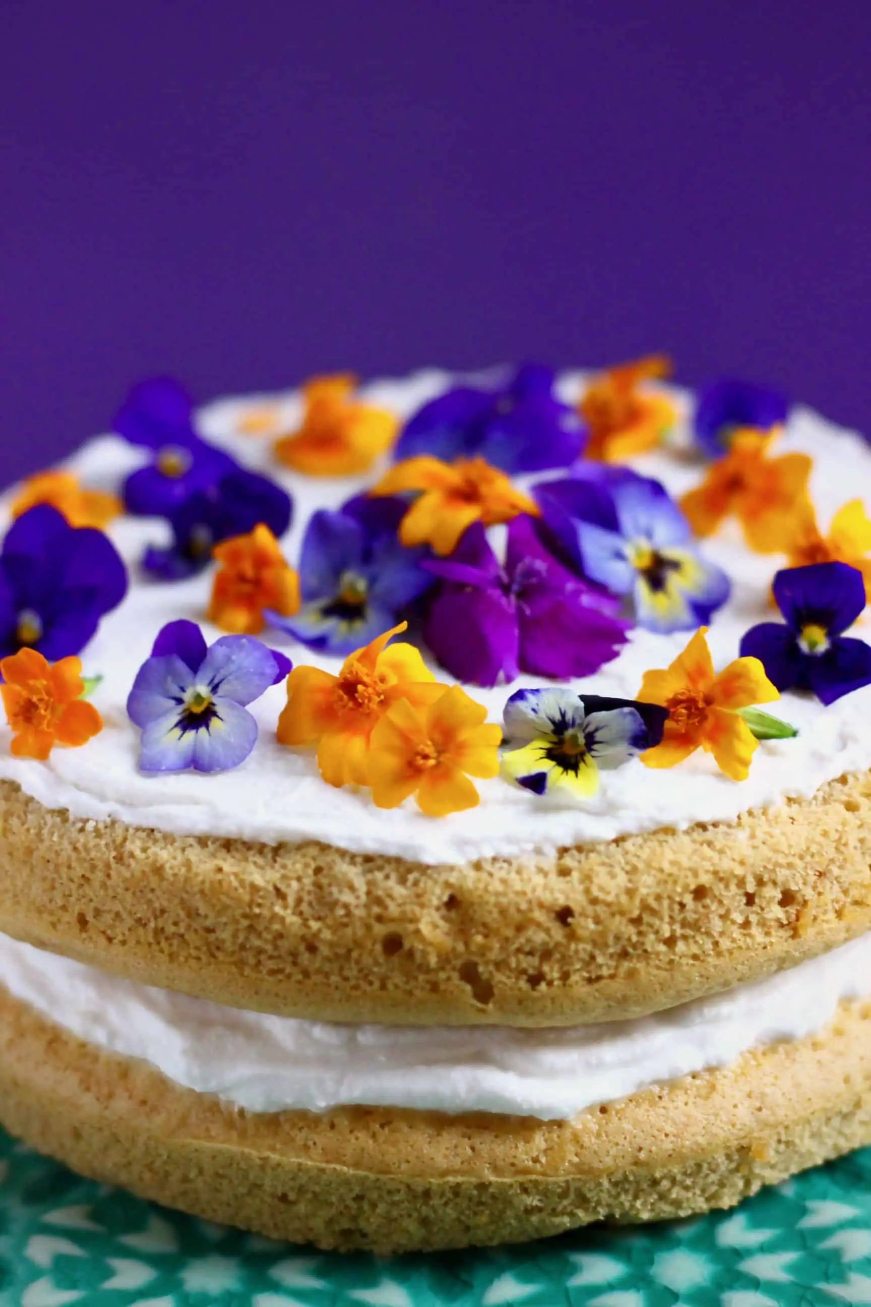 A pumpkin sponge sandwiched with white frosting topped with purple and orange flowers on a green cake stand against a purple background