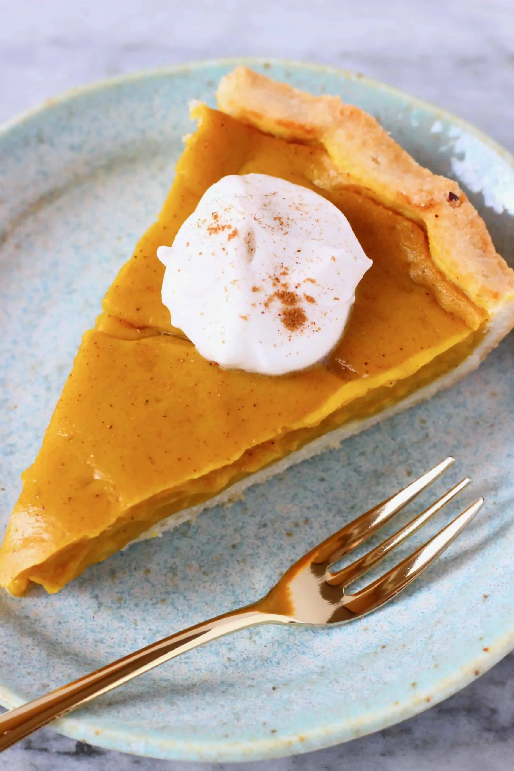 A slice of pumpkin pie topped with cream and sprinkled with cinnamon on a blue plate with a gold fork